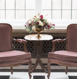 Vintage purple velvet armchairs with wooden arms. In between the chairs is a dark wood vintage side table with flowers in a vase. Naturally backlit by a large window.