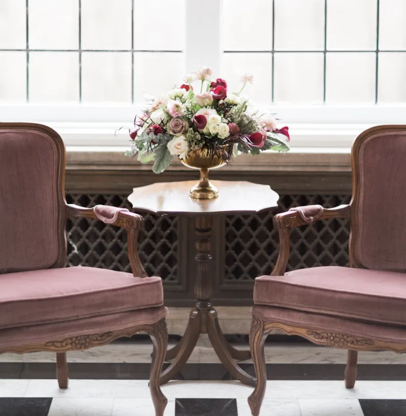Purcle armchairs with vintage dark wood side table. WEBP Vintage purple velvet armchairs with wooden arms. In between the chairs is a dark wood vintage side table with flowers in a vase. Naturally backlit by a large window.