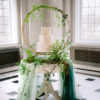 Three tiered wedding cake sitting on ornate vintage round table with a big circle decor and greenery