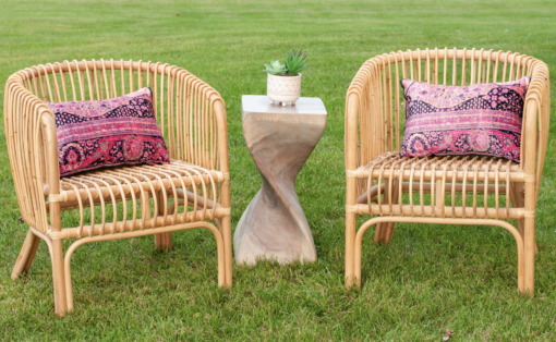 Two rattan natural wood colored chairs with pink pillows on each. Curved side table in between with plant in white vase. Outside on grass.