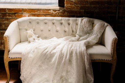 Cream loveseat with tufted button back and wooden arms and legs. Wedding dress laying on loveseat. Against a brick wall.