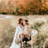 Couple sitting on an oversized rattan chair in a field. Fall trees in the background.