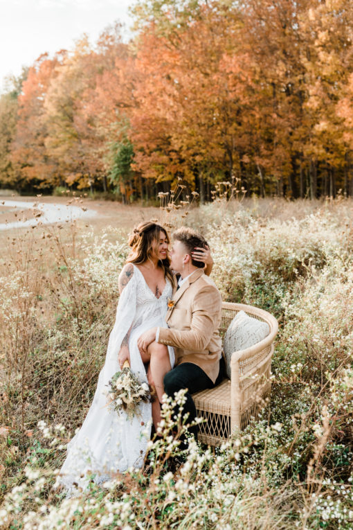 Couple sitting on an oversized rattan chair in a field. Fall trees in the background.