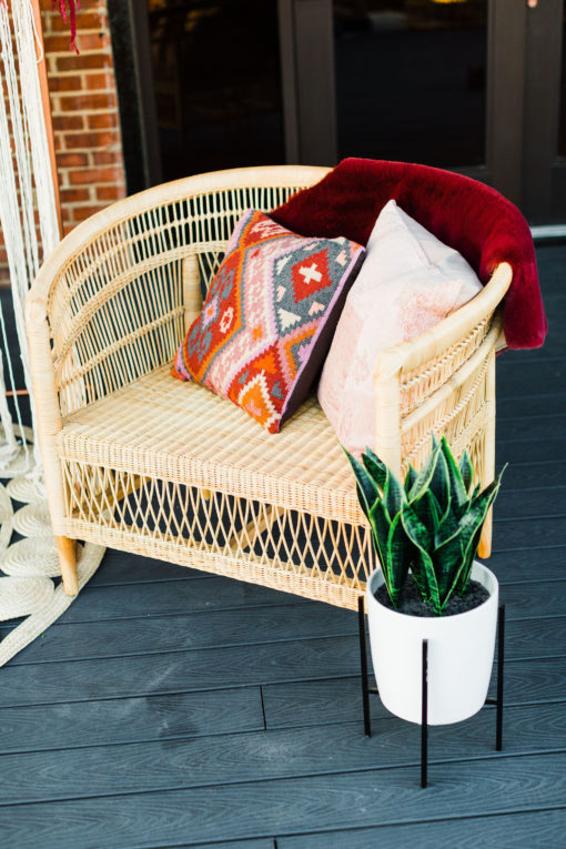 Oversided rattan chair on a deck. Two pillows and a throw over the edge. Snake plant in a white vase next to the chair.