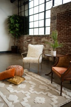 A bohemian-chic lounge setup from a styled shoot in downtown Indianapolis, featuring a white faux-fur gold-framed armchair and a vintage velvet cane chair. The scene is set against an industrial brick wall and large factory windows, complete with a patterned cream rug, a leather Moroccan pouf, and terracotta accent pillows, perfect for wedding or event inspiration.