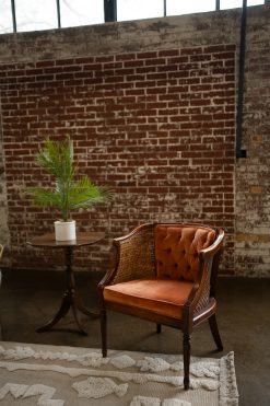 A vintage orange velvet and cane-sided chair sits next to a small wooden side table with a potted palm plant, on a patterned cream rug, against an exposed red brick wall in an industrial loft in downtown Indianapolis, Indiana, perfect for a wedding, reception, or corporate event.