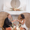 Young diverse couple sitting in rounded high back peacock chairs at a boho themed sweetheart table at wedding reception in Carmel, Indiana. Florals on the table and a round clock behind.