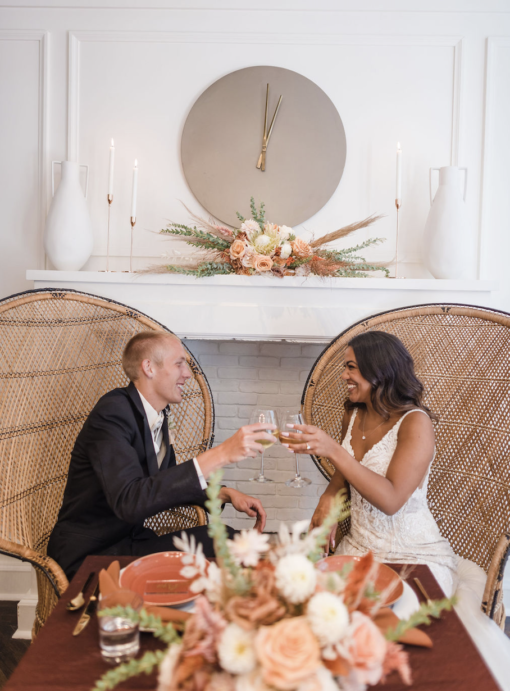 Young diverse couple sitting in rounded high back peacock chairs at a boho themed sweetheart table at wedding reception in Carmel, Indiana. Florals on the table and a round clock behind.