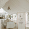 White wedding reception with a decorated table in the background and a tall copper sign in the foreground with a banner of the couple's names