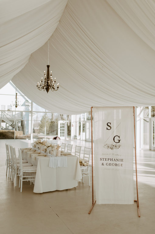 White wedding reception with a decorated table in the background and a tall copper sign in the foreground with a banner of the couple's names