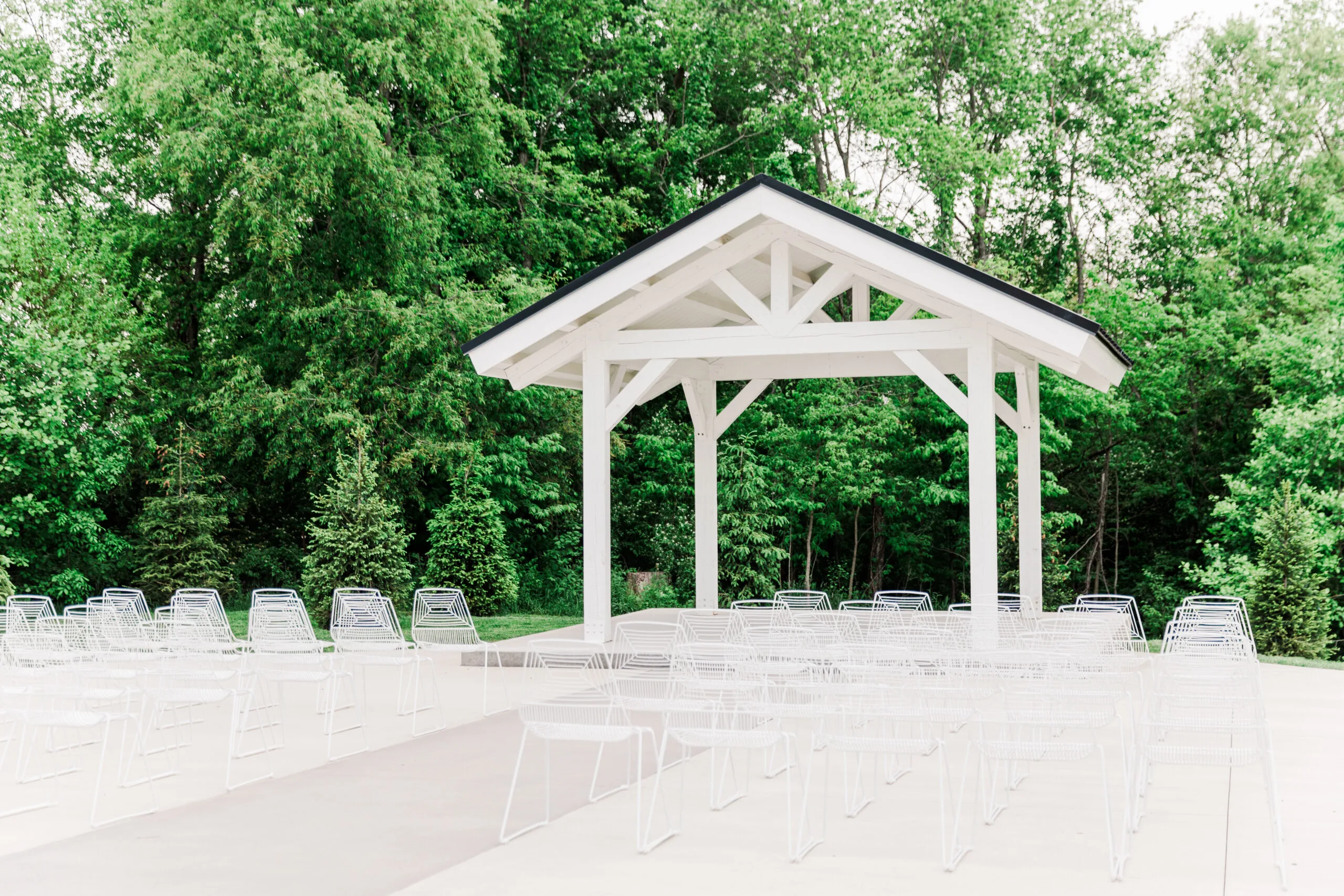 Outdoor wedding ceremony with white classic chairs WEBP White wire chairs set up for an outdoor wedding ceremony. Green trees in background with a small covered structure