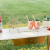 Mid-century modern desk with marble top with inlaid parallel gold lines. Gold legs and desk drawer. Bottles of upscale soda are on the table along with cookies, a decorative star, a plant, and two copper mule mugs.