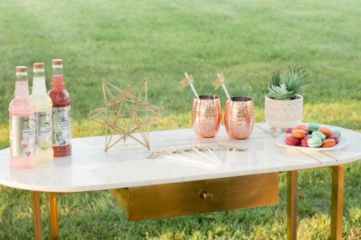 Mid-century modern desk with marble top with inlaid parallel gold lines. Gold legs and desk drawer. Bottles of upscale soda are on the table along with cookies, a decorative star, a plant, and two copper mule mugs.