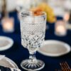 Etched glass goblet on a pedestal against a dark blue linen. White plates in the background with florals