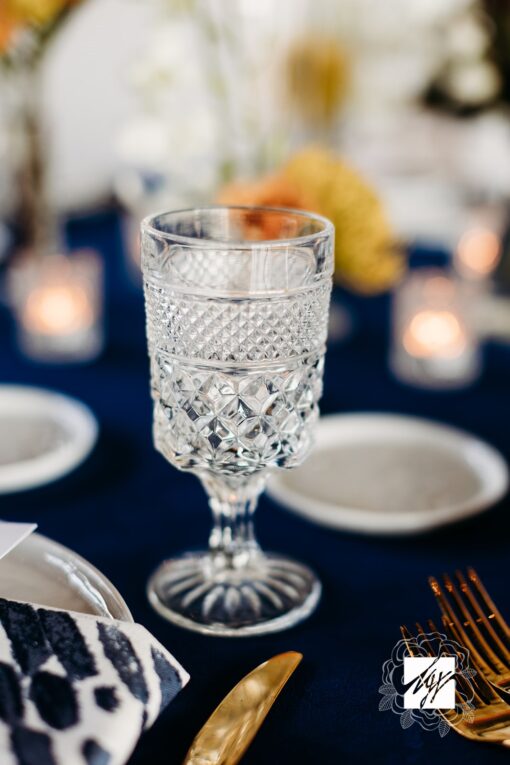 Etched glass goblet on a pedestal against a dark blue linen. White plates in the background with florals