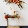 Wooden bar cart with brass grame against a white wall. Orange florals on the back. White three tiered cake on the card.