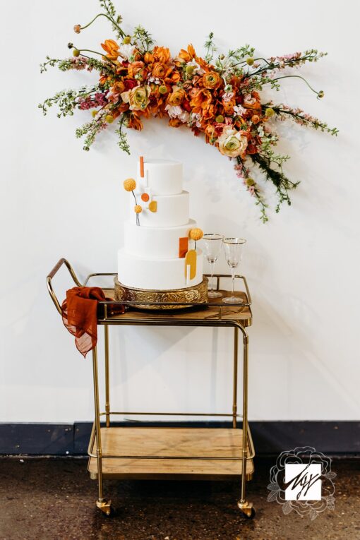 Wooden bar cart with brass grame against a white wall. Orange florals on the back. White three tiered cake on the card.