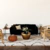 Lounge setting against a white wall. Black sofa with gold coffee and side table. Two rattan chairs sitting on a black and white shaggy rug. Brown leather pouf.