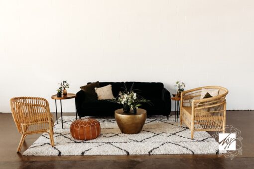 Lounge setting against a white wall. Black sofa with gold coffee and side table. Two rattan chairs sitting on a black and white shaggy rug. Brown leather pouf.