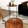 Brown leather pouf sitting on a black and white shaggy rug with black sofa and side table in the background