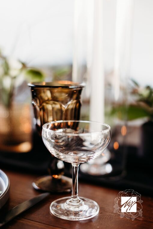 Clear coupe with a wide rim on a table with brown goblet and white candle behind.