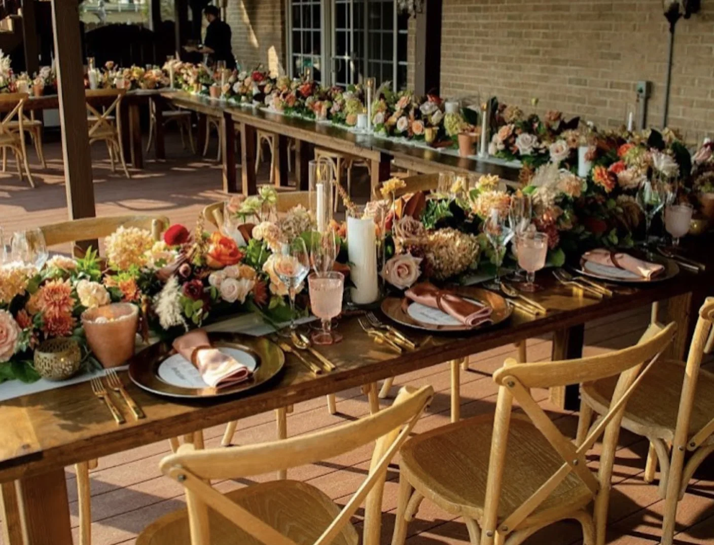 Vineyard x-back chairs around wooden farm tables in outdoor dinner WEBP Light wood x-back chairs in a U shape head table with wooden farm tables and lovely florals and tablescape at an outdoor wedding reception dinner.