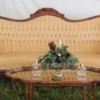 Vintage curved sofa in a pale yellow with dark wood edging. Wooden coffee table with glass top and florals. Outdoors on grass in a lounge seating area.