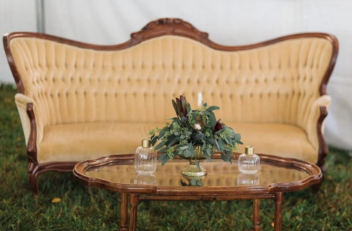 Vintage curved sofa in a pale yellow with dark wood edging. Wooden coffee table with glass top and florals. Outdoors on grass in a lounge seating area.