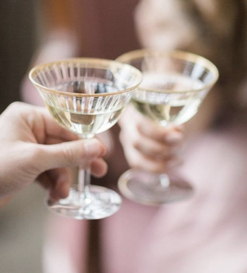 Close up on two vintage coupes with gold rims being held by 2 people in a toast.