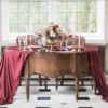 Ornate vintage dark wooden crescent shaped desk. Used as a sweetheart table in a wedding reception. Desk draped with a maroon runner, brass candlesticks, and two vintage arm chairs in the back. Natural light from window behind.