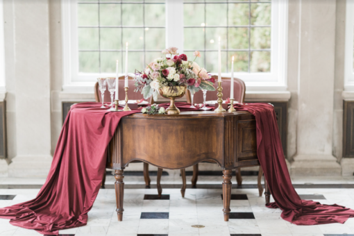 Ornate vintage dark wooden crescent shaped desk. Used as a sweetheart table in a wedding reception. Desk draped with a maroon runner, brass candlesticks, and two vintage arm chairs in the back. Natural light from window behind.