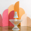 Bright colors on an arched and curved paneled backdrop. Tallest panel is tangerine, middle is hot pink, smallest is blush. Against a white wall. White table with wedding cake placed in front.