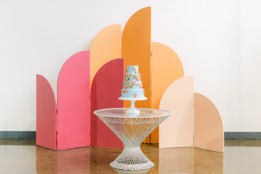 Bright colors on an arched and curved paneled backdrop. Tallest panel is tangerine, middle is hot pink, smallest is blush. Against a white wall. White table with wedding cake placed in front.