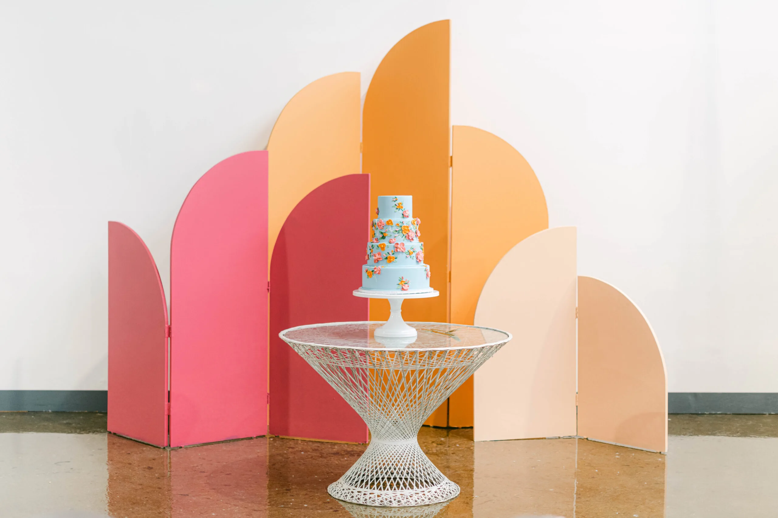 Ezra curved backdrop with wedding cake WEBP Bright colors on an arched and curved paneled backdrop. Tallest panel is tangerine, middle is hot pink, smallest is blush. Against a white wall. White table with wedding cake placed in front.