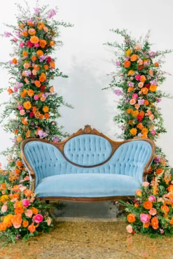 Vintage curved blue velvet sofa set in a backdrop of two floral pillars with bright orange, pink, and green. White background and concrete floor