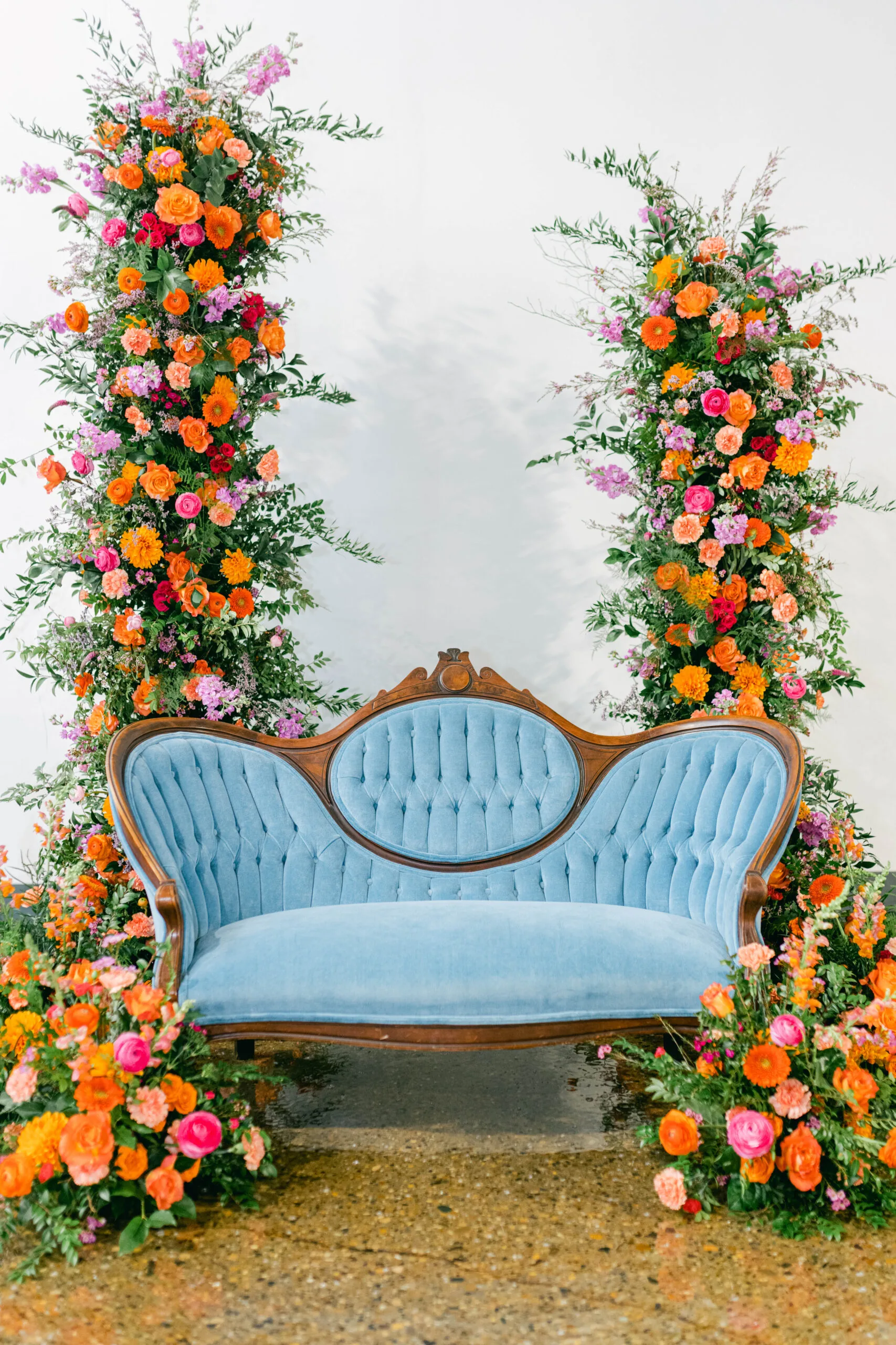 Lamere Loveseat WEBP Vintage curved blue velvet sofa set in a backdrop of two floral pillars with bright orange, pink, and green. White background and concrete floor