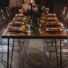 Side view of a wooden farm table with black hairpin legs. Decorated for a wedding reception with clear ghost chairs and amber tablescape decor.