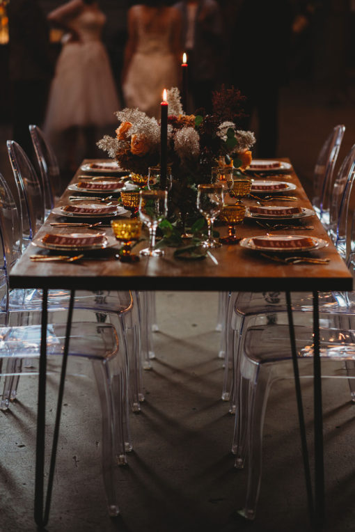 Side view of a wooden farm table with black hairpin legs. Decorated for a wedding reception with clear ghost chairs and amber tablescape decor.