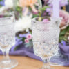 Matching cut glass goblets with purple florals and candlesticks in background at a wedding reception tablescape