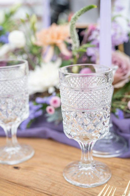 Matching cut glass goblets with purple florals and candlesticks in background at a wedding reception tablescape