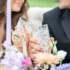 Bridal couple with crystal glasses in a toast. Florals in the foreground, blurred couple in the back. Image focused on the glasses