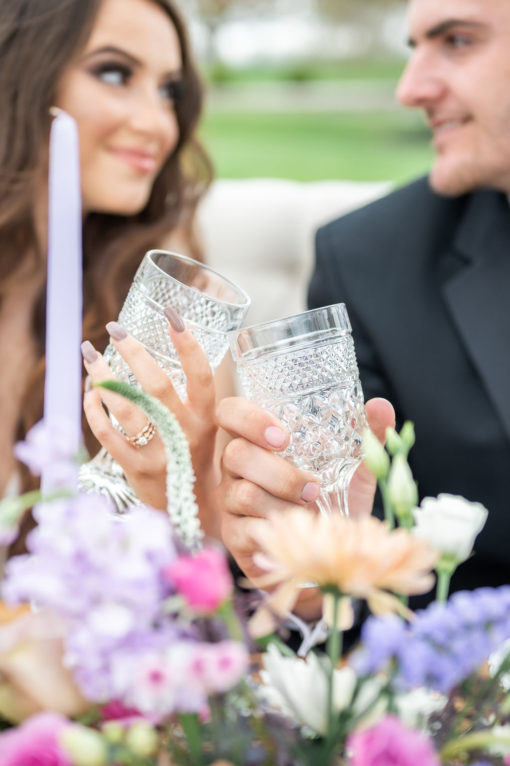 Bridal couple with crystal glasses in a toast. Florals in the foreground, blurred couple in the back. Image focused on the glasses