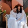 Wives posing with the Ezra backdrop. Both women wearing white dresses, sitting in oversized rattan chair.