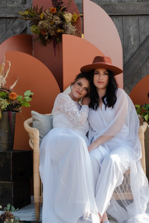 Wives posing with the Ezra backdrop. Both women wearing white dresses, sitting in oversized rattan chair.