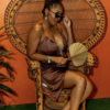 Woman posing while seating in a vintage peacock chair. She's pulling down her sunglasses and looking at the camera. Orange wall in the background.