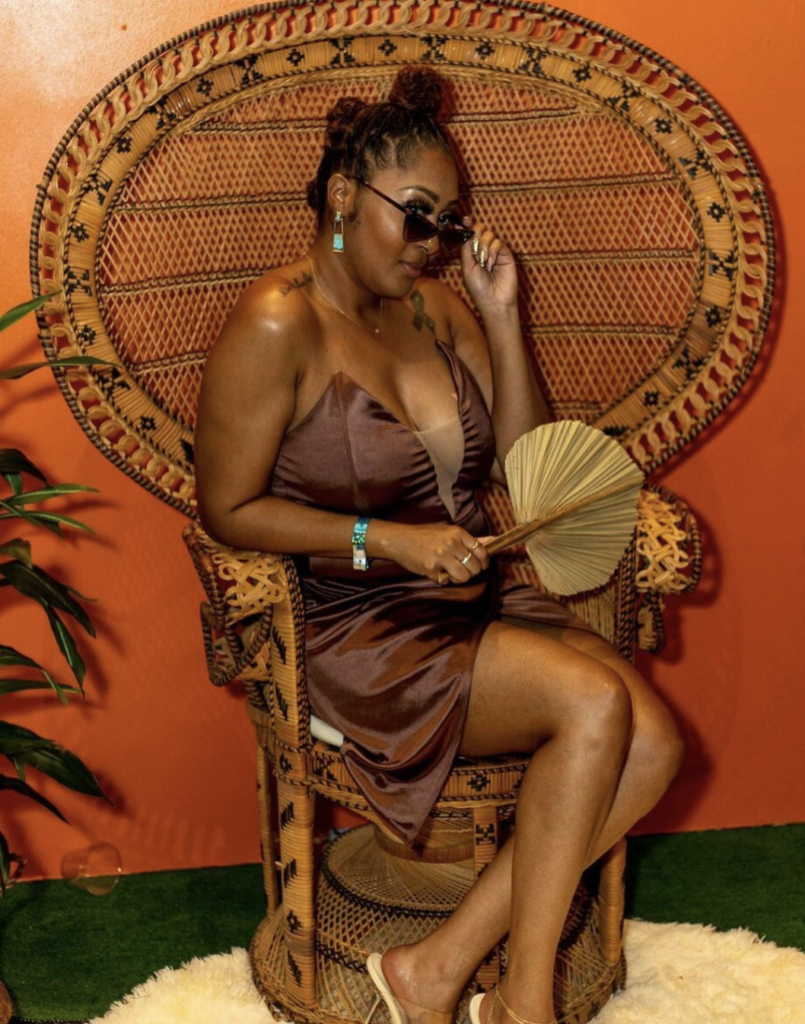 Woman posing while seating in a vintage peacock chair. She's pulling down her sunglasses and looking at the camera. Orange wall in the background.