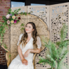 Woman in a white dress sitting on a peacock chair with florals. Wooden backdrop behind. Large plant on the right.