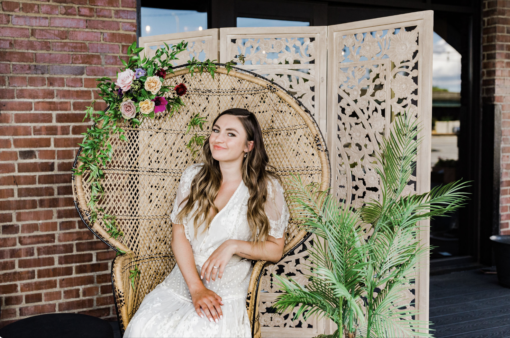 Woman in a white dress sitting on a peacock chair with florals. Wooden backdrop behind. Large plant on the right.