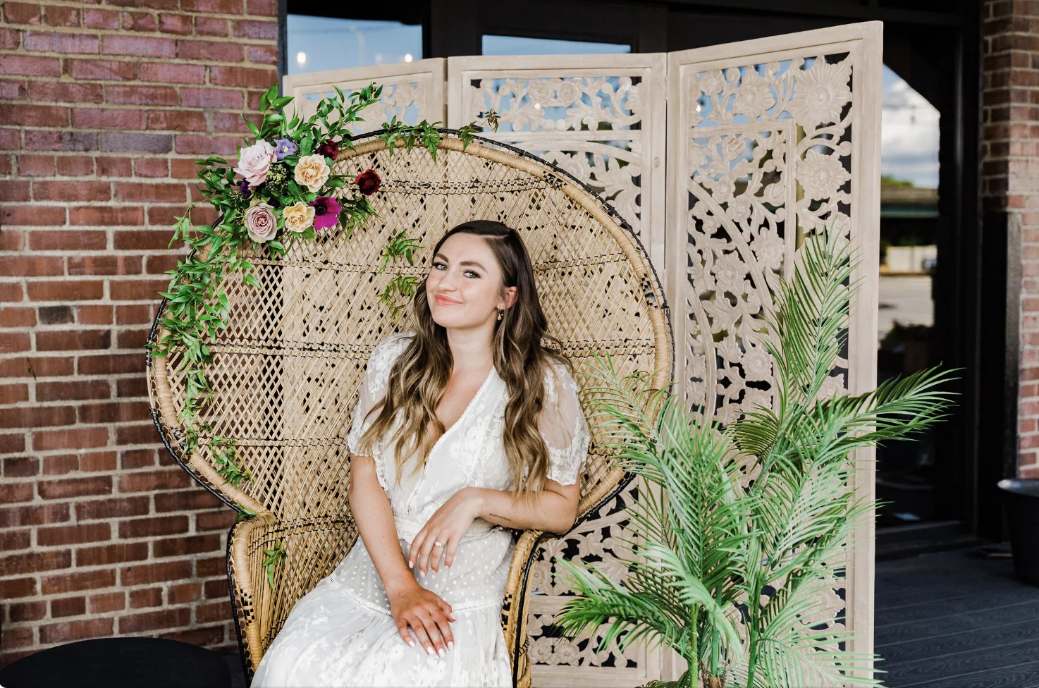Woman sitting on peacock chair WEBP Woman in a white dress sitting on a peacock chair with florals. Wooden backdrop behind. Large plant on the right.