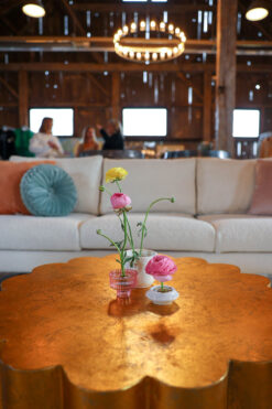 Gold metal coffee table with scallop edges in the foreground and a white soft in the background against a wooden barn wall.